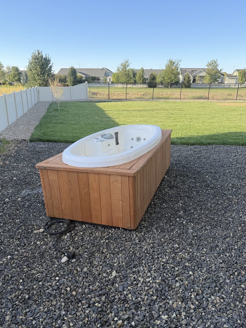 Cedar-sided oval hot tub sitting on a gravel pad in a Meridian, Idaho backyard before HaulWorks removed it
