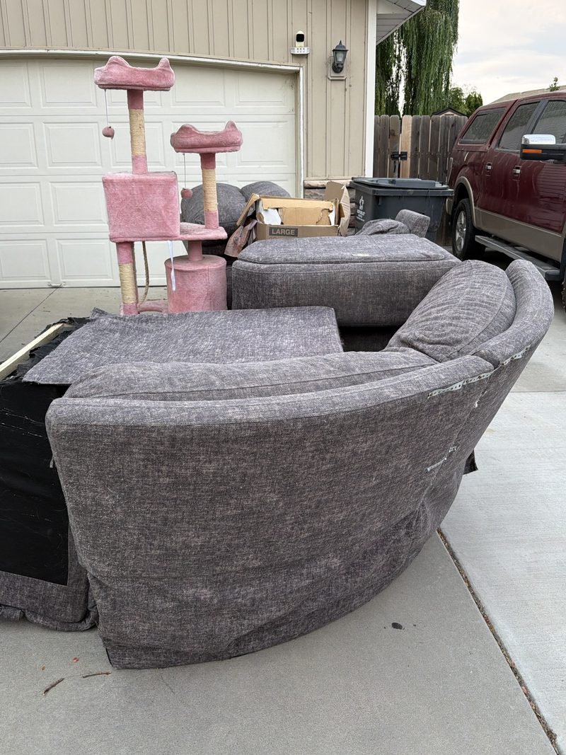 Grey sectional sofa, ottoman, cushions and a cat tree stacked on a Meridian driveway before HaulWorks furniture removal